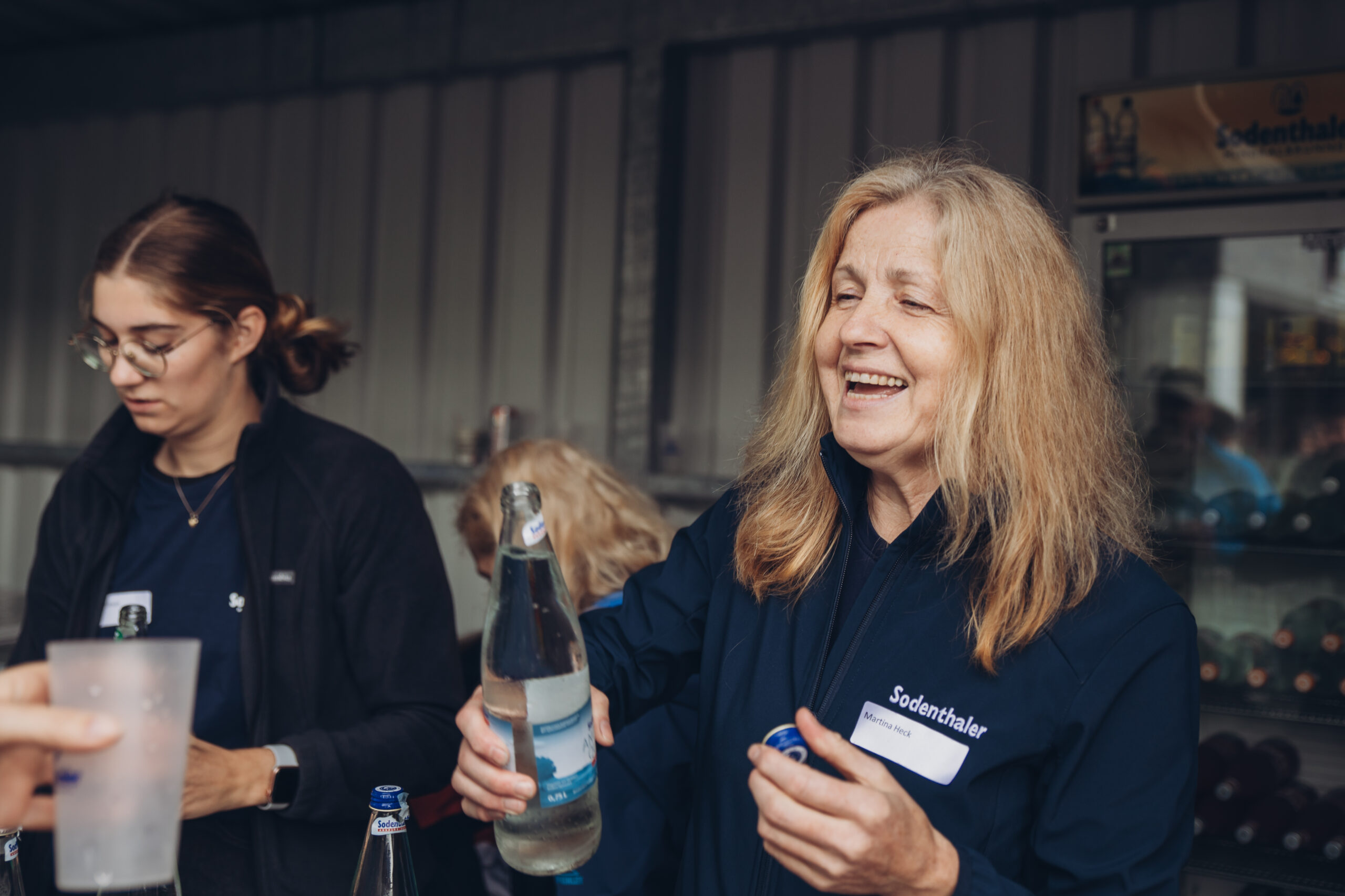 Eine lachende Frau schenkt Wasser aus einer Glasflasche aus, während eine weitere Frau im Hintergrund arbeitet. Beide tragen Kleidung mit dem Logo „Sodenthaler“.