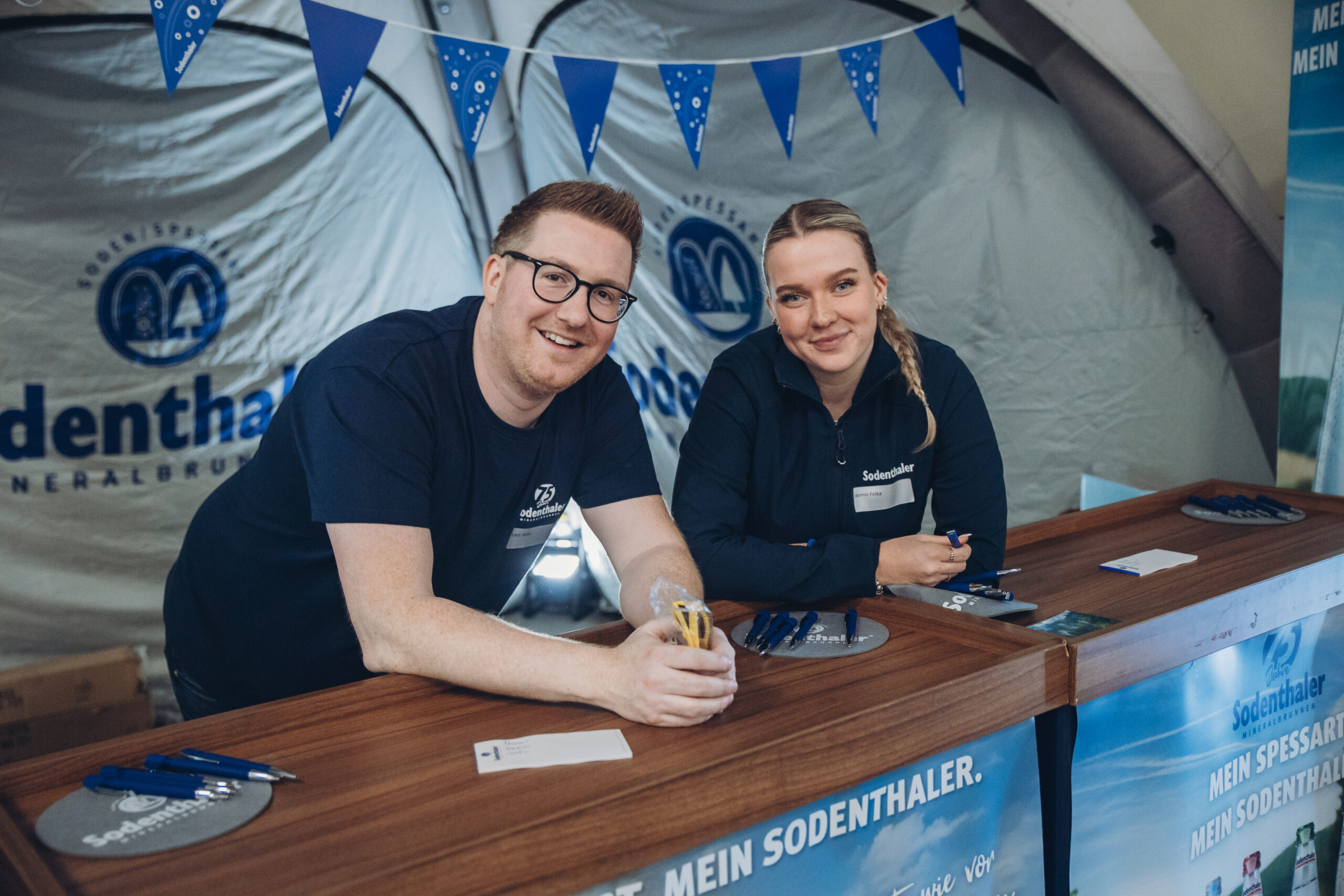 Ein Mann und eine Frau sitzen an einem Infostand mit Sodenthaler-Branding. Sie lächeln freundlich, der Hintergrund zeigt ein Zelt mit Logos.