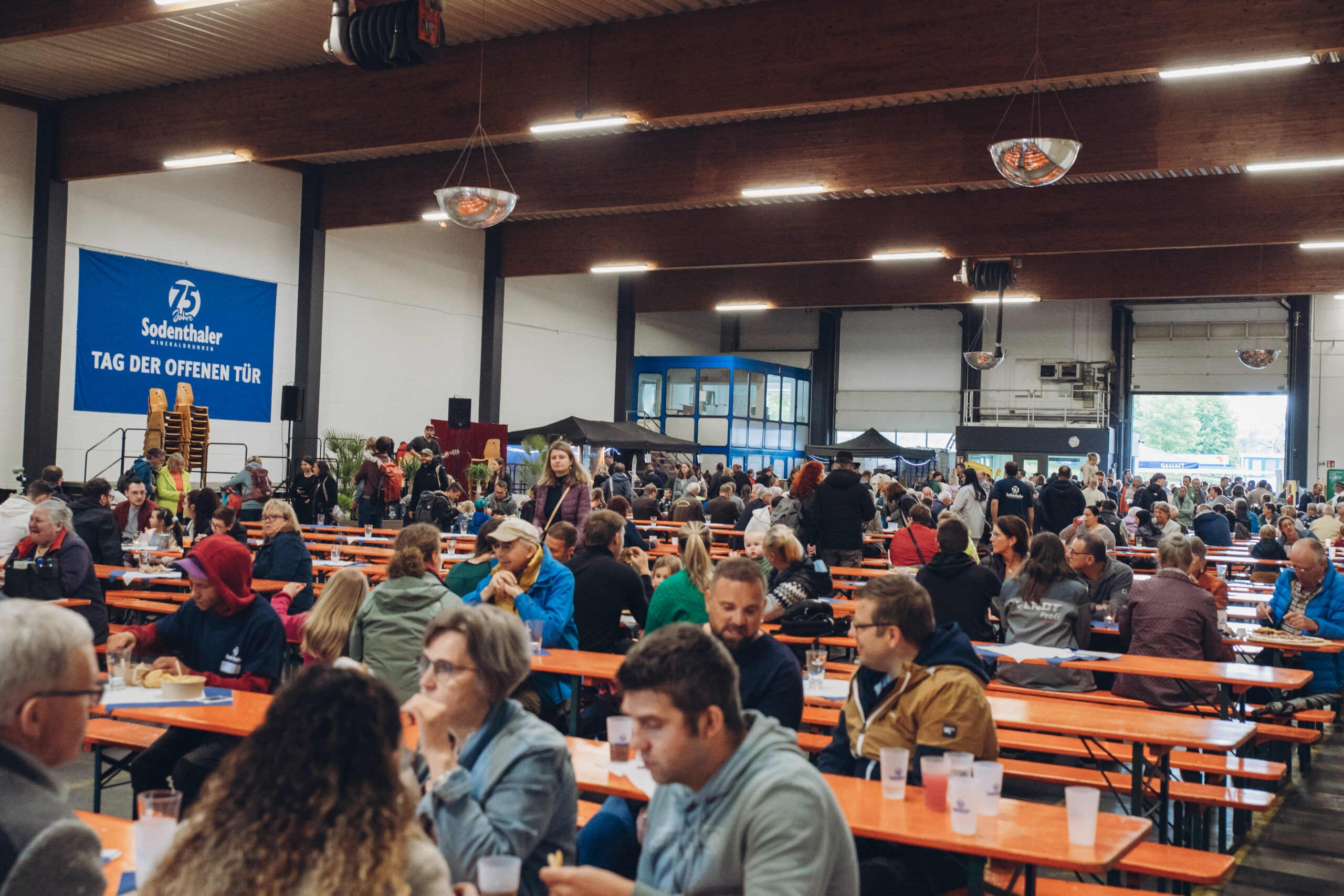 Viele Besucher sitzen an langen Tischen in einer großen Halle, dekoriert mit einem „Tag der offenen Tür“-Banner von Sodenthaler.
