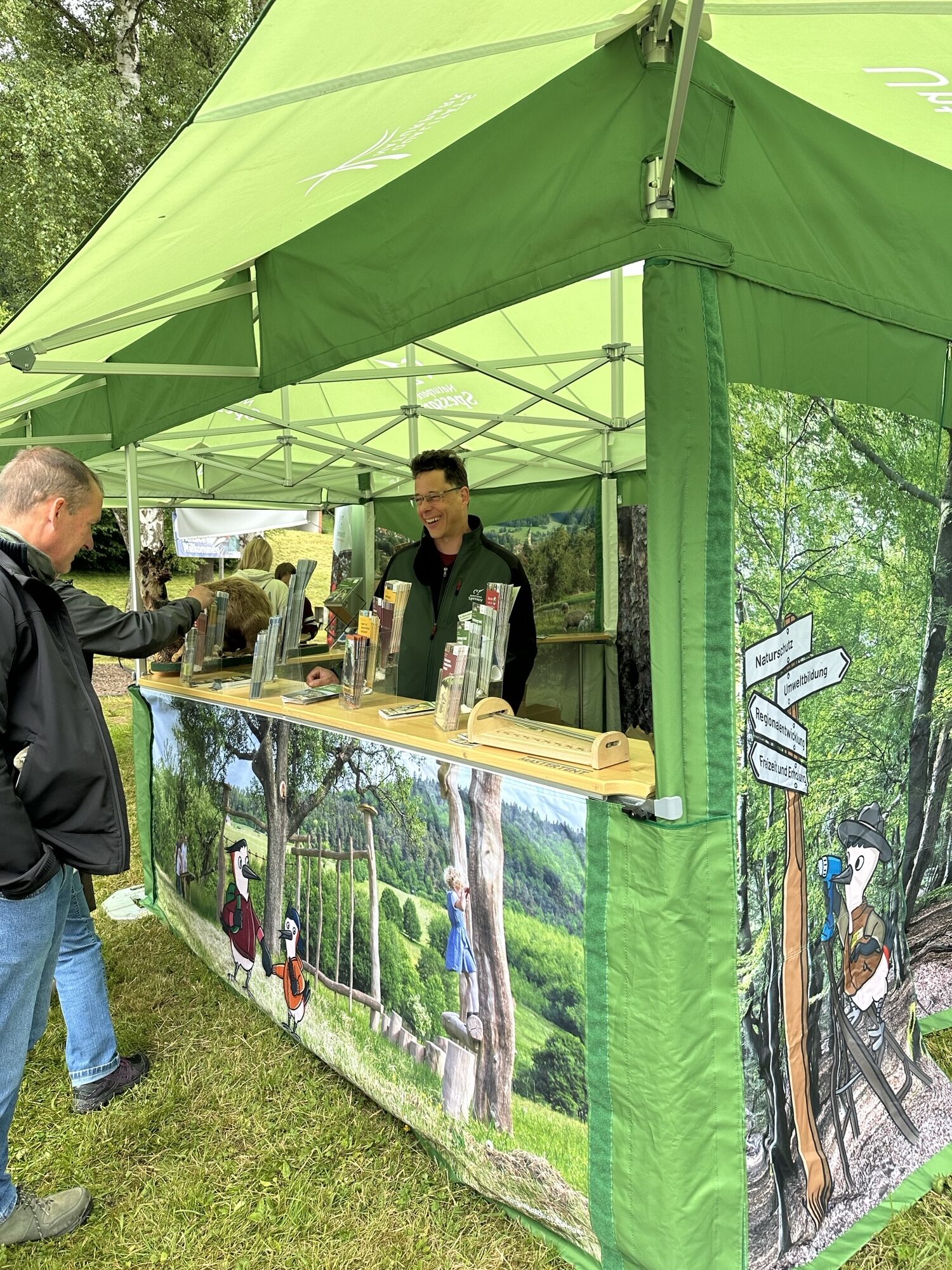 Ein Mitarbeiter informiert Besucher an einem grünen Stand mit Naturmotiven über Wanderwege und Freizeitangebote im Spessart.