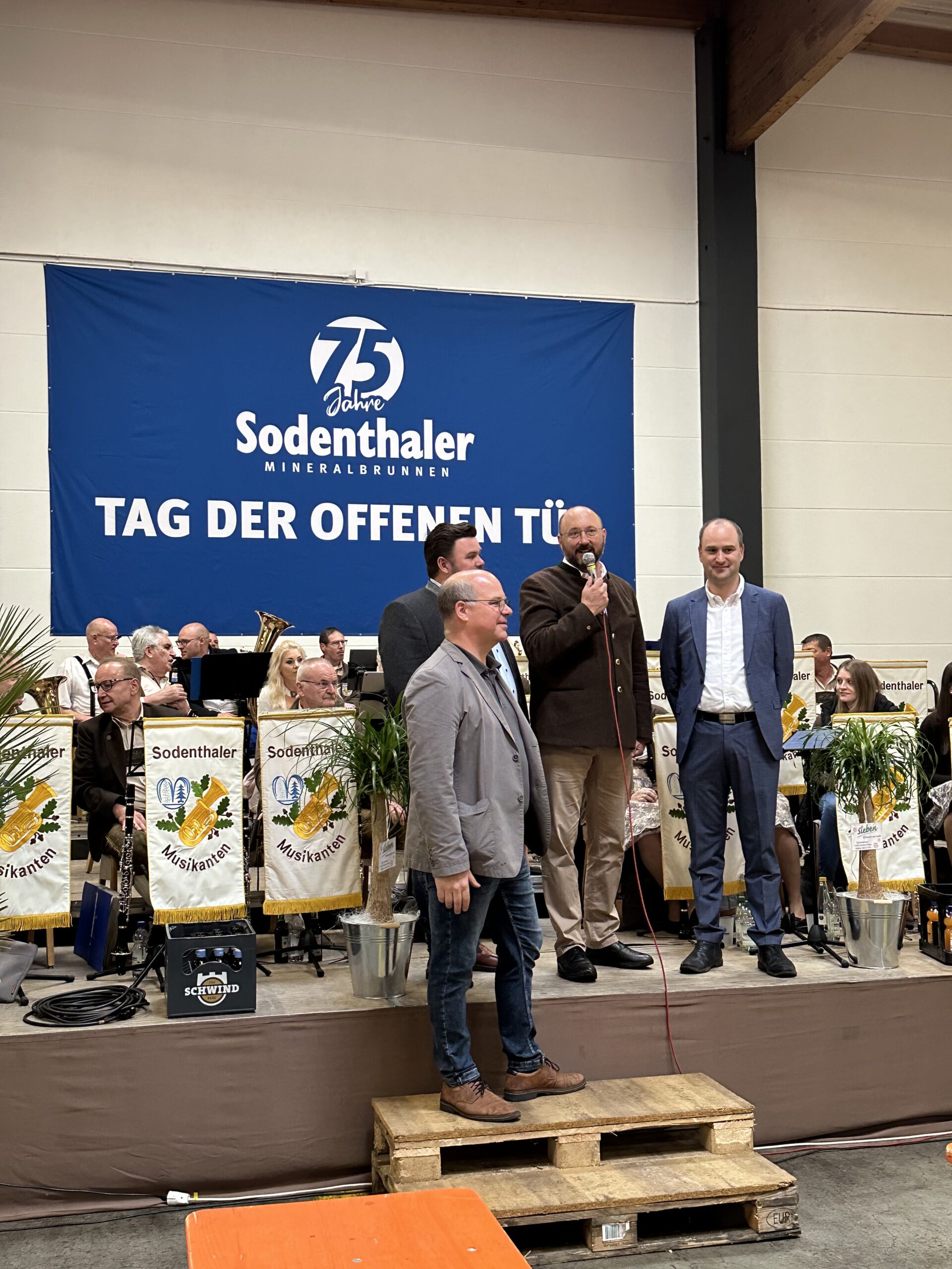 Mehrere Männer halten eine Ansprache auf einer Bühne vor einem Banner zum 75-jährigen Jubiläum von Sodenthaler, begleitet von einer Blaskapelle.