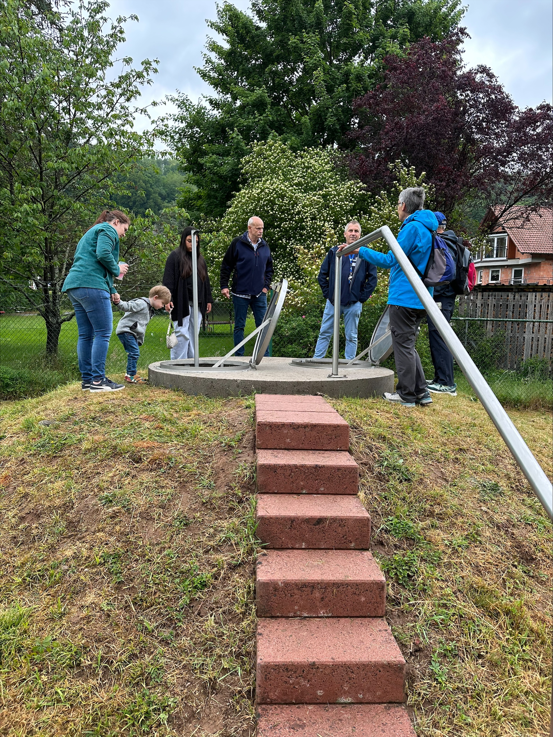 Menschen sammeln sich um einen Trinkbrunnen auf einer kleinen Anhöhe im Grünen.